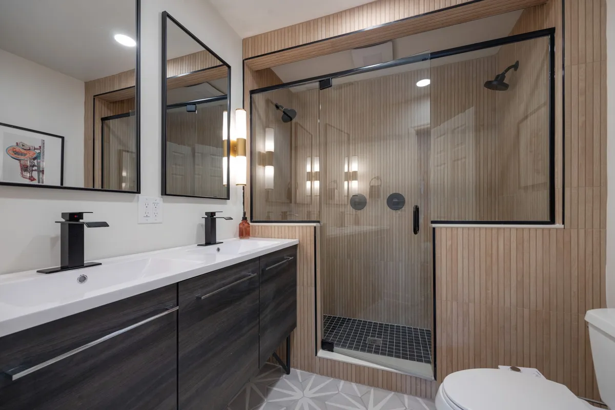 A serene, contemporary ensuite bathroom featuring a sleek vanity with dual sinks, framed mirrors, a glass-enclosed shower, and modern light fixtures. The ambiance is relaxing, ideal for self-care and winding down after a day out.