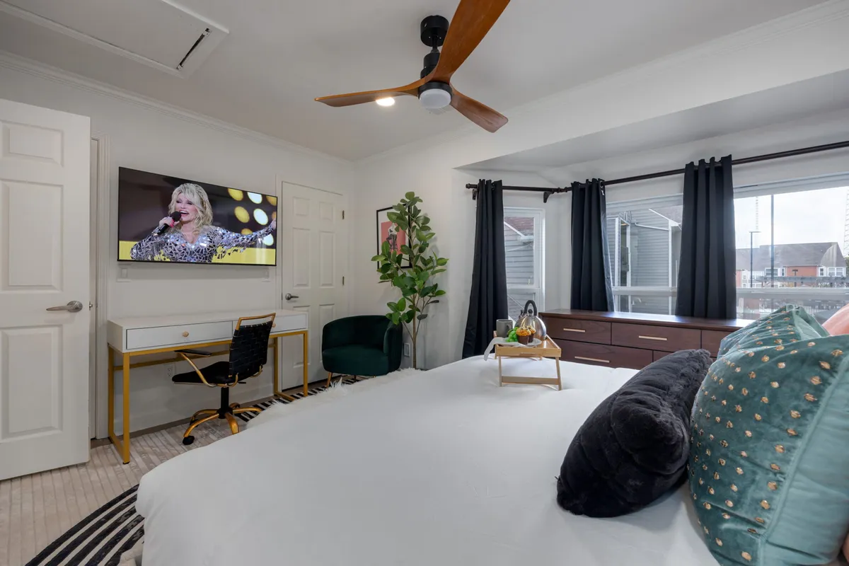 A well-appointed bedroom featuring a cozy bed with decorative pillows, a wall-mounted smart TV displaying a lively music performance, a decorative plant, and a dedicated desk with a chair, ideal for work or study. The room is designed for both relaxation and productivity.