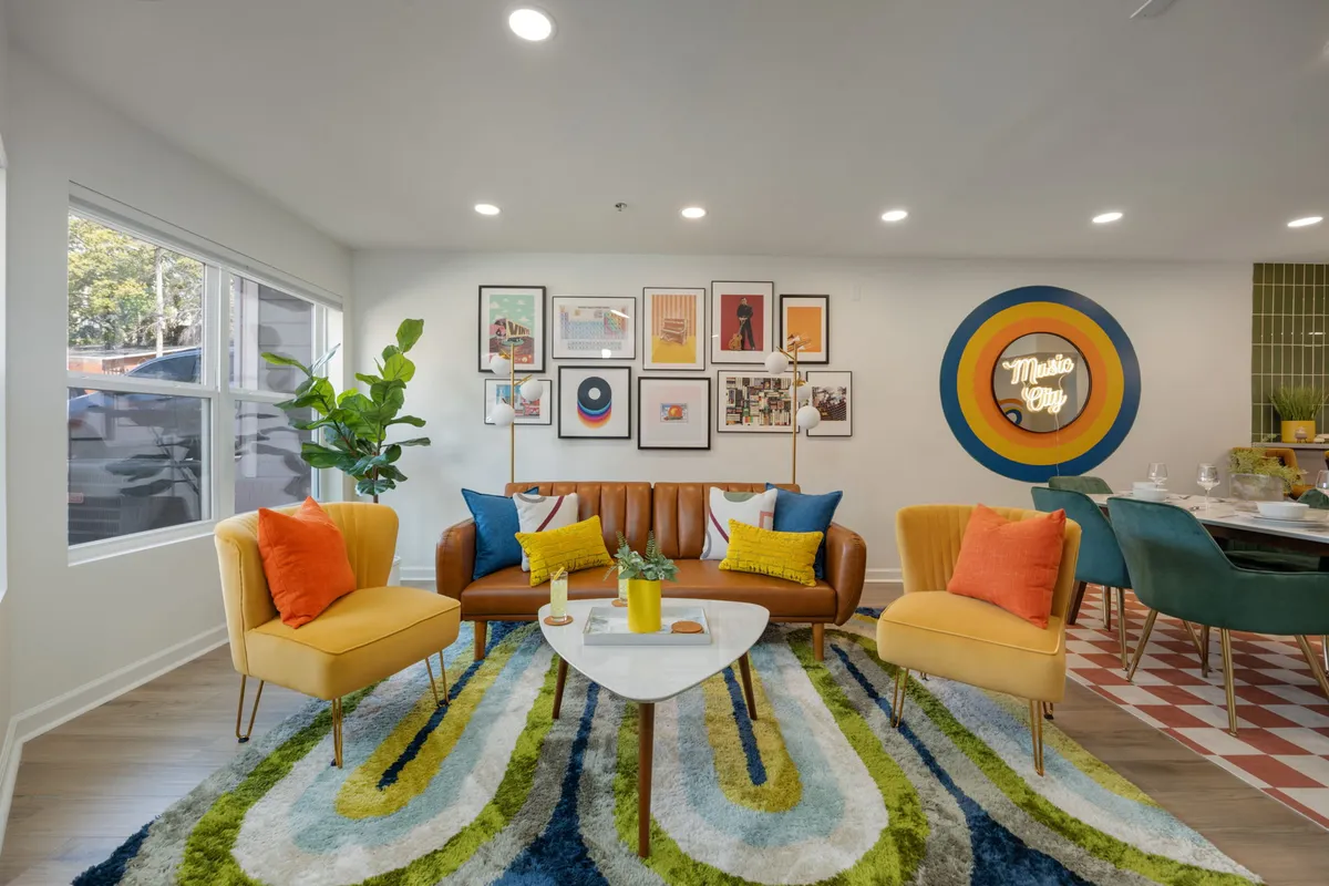 A playful, open-plan living room featuring a colorful couch, a gallery art wall, a unique patterned rug, and music-themed wall decor. The space is bright and welcoming, ideal for socializing, relaxing, or hosting a lively board game night.