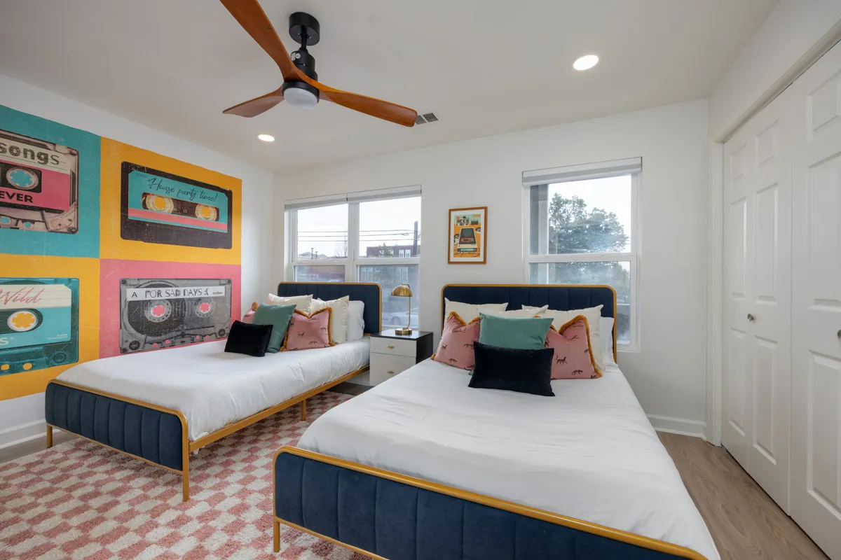A cheerful bedroom with two full beds, each dressed in colorful pillows and bedding. A large, vibrant mural graces the wall, while sunlight pours in through big windows, creating a bright, playful environment ideal for families or friends. The overall mood is cozy and inviting.