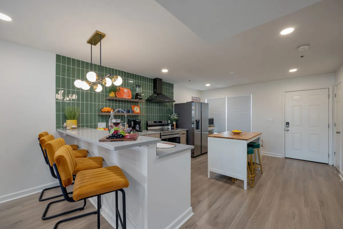 A warm, contemporary kitchen with a green tiled backsplash, stylish modern lights, a large island with bright yellow stools, and a fresh fruit arrangement. The space feels inviting for cooking, casual dining, and socializing.