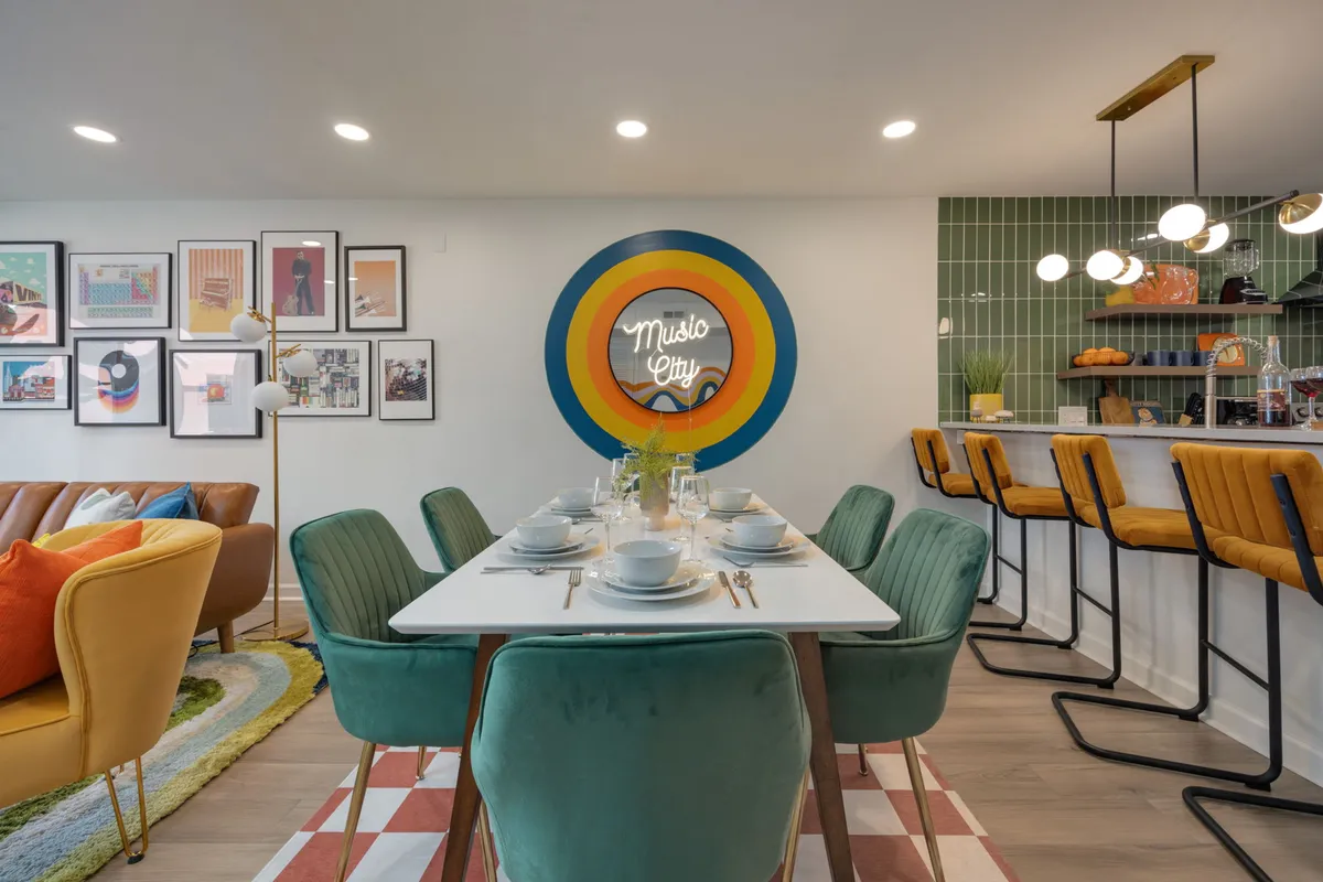 A stylish dining area with a table set for six, colorful wall art, a neon 'Music City' sign, and an adjacent bar seating area. The mood is lively and the scene is ready for both family meals and festive celebrations.