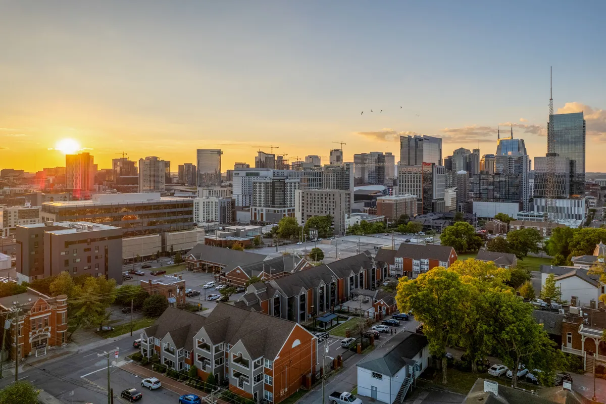 A scenic view over a neighborhood with lush trees, residential buildings, and Nashville’s skyline in the background, all framed by the warm glow of sunset.