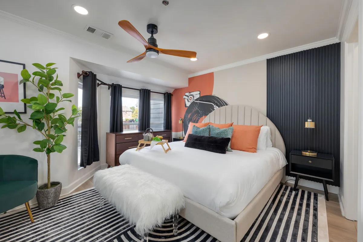 A bright, stylish bedroom with a plush bed dressed in inviting bedding, vibrant wall art above the headboard, a green plant for a touch of nature, and a bold striped rug underfoot. Natural light streams through the windows, creating a cozy, welcoming atmosphere perfect for relaxation.