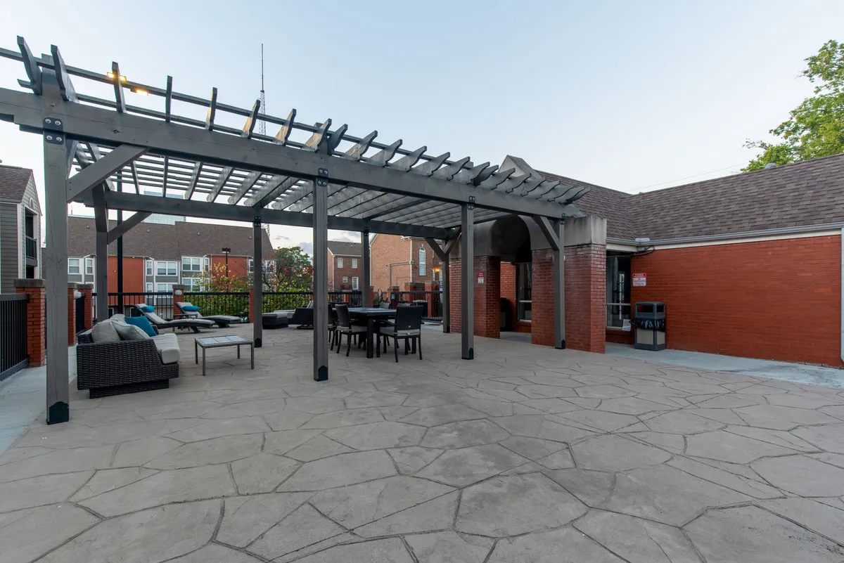 A pergola shades an outdoor seating area with a dining table, creating a cozy space ideal for al fresco meals or evening drinks. The surrounding architecture suggests a lively, communal urban setting.